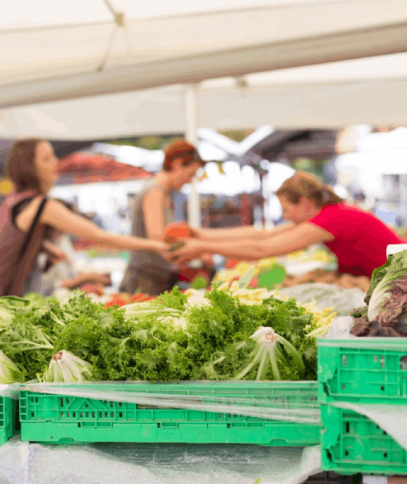 Fresh vegetables on the counter of a farmers market with customers in the background<br />